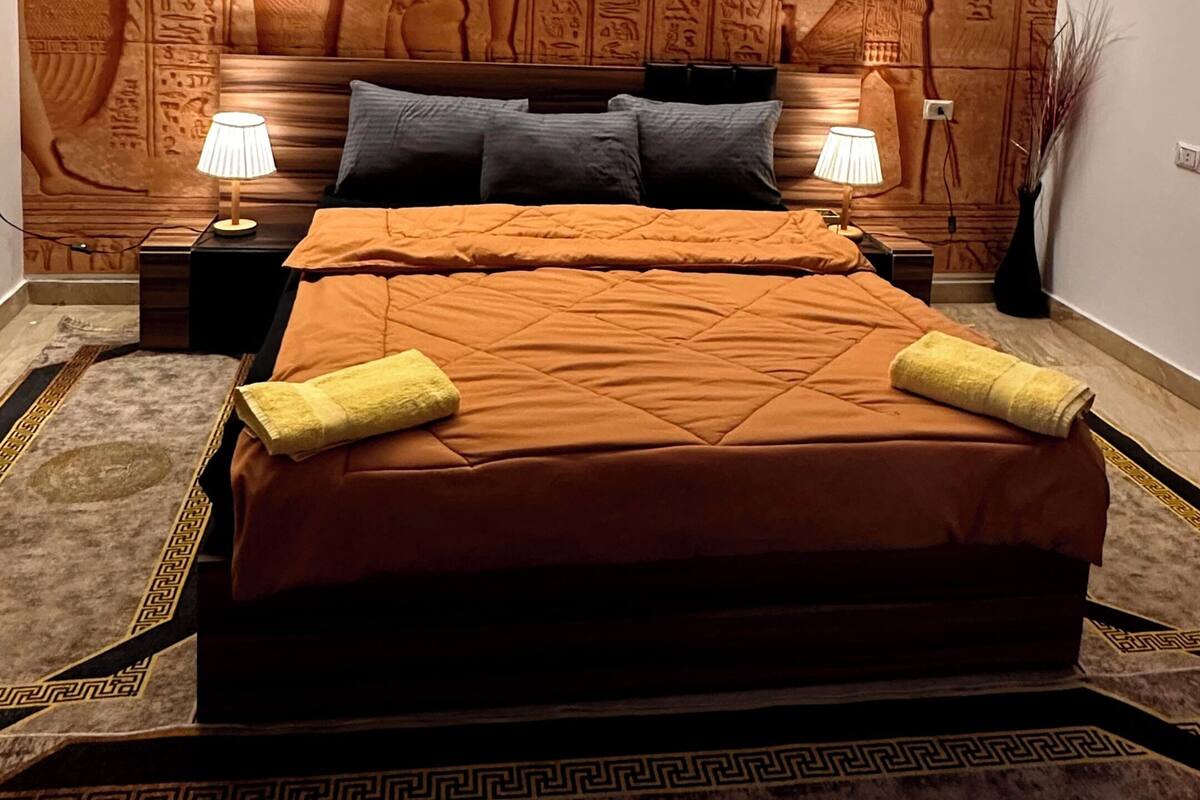 A bedroom with an Egyptian-themed wall mural, a bed with a patterned bedspread, two bedside lamps, and a decorative vase.