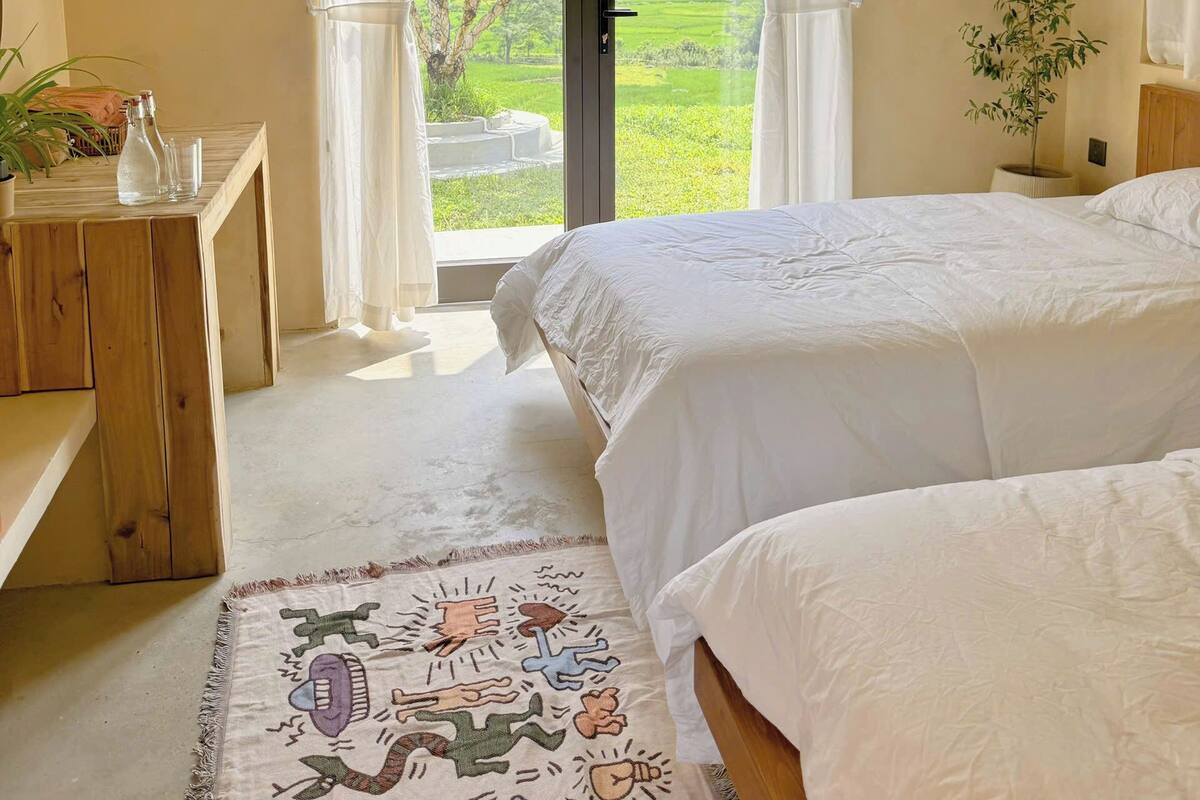 A bedroom with a bed, a bedside table, a rug, and a view of a green landscape through a glass door.