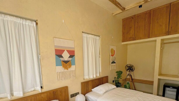 Ein Hotelzimmer mit zwei Betten, Holz-Kopfteilen, weißen Vorhängen, einem Deckenventilator und einer kleinen Pflanze.