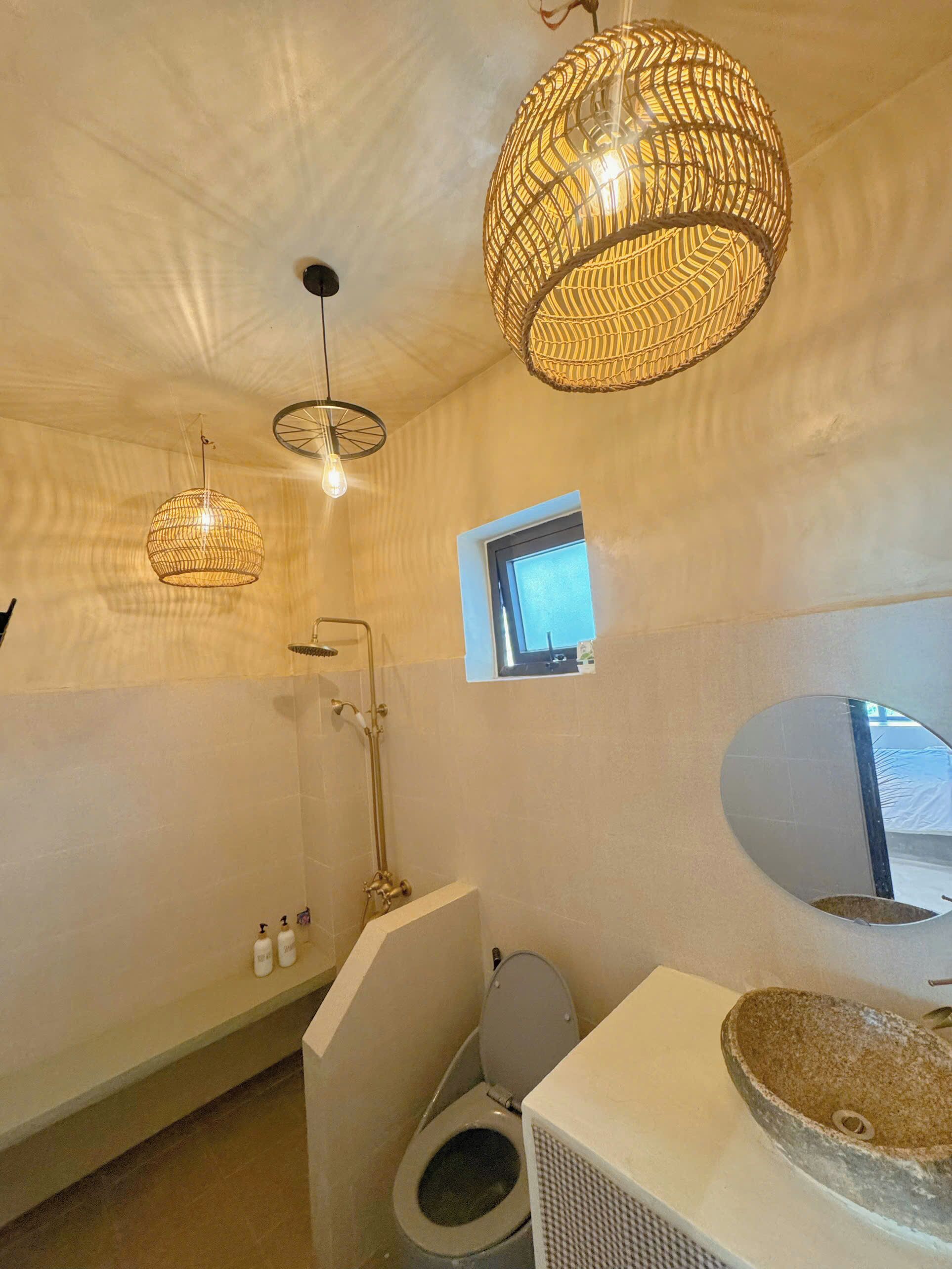 Een badkamer met een toilet, een douche en een wastafel met een stenen wasbak. Er zijn twee hanglampen en een klein raam.