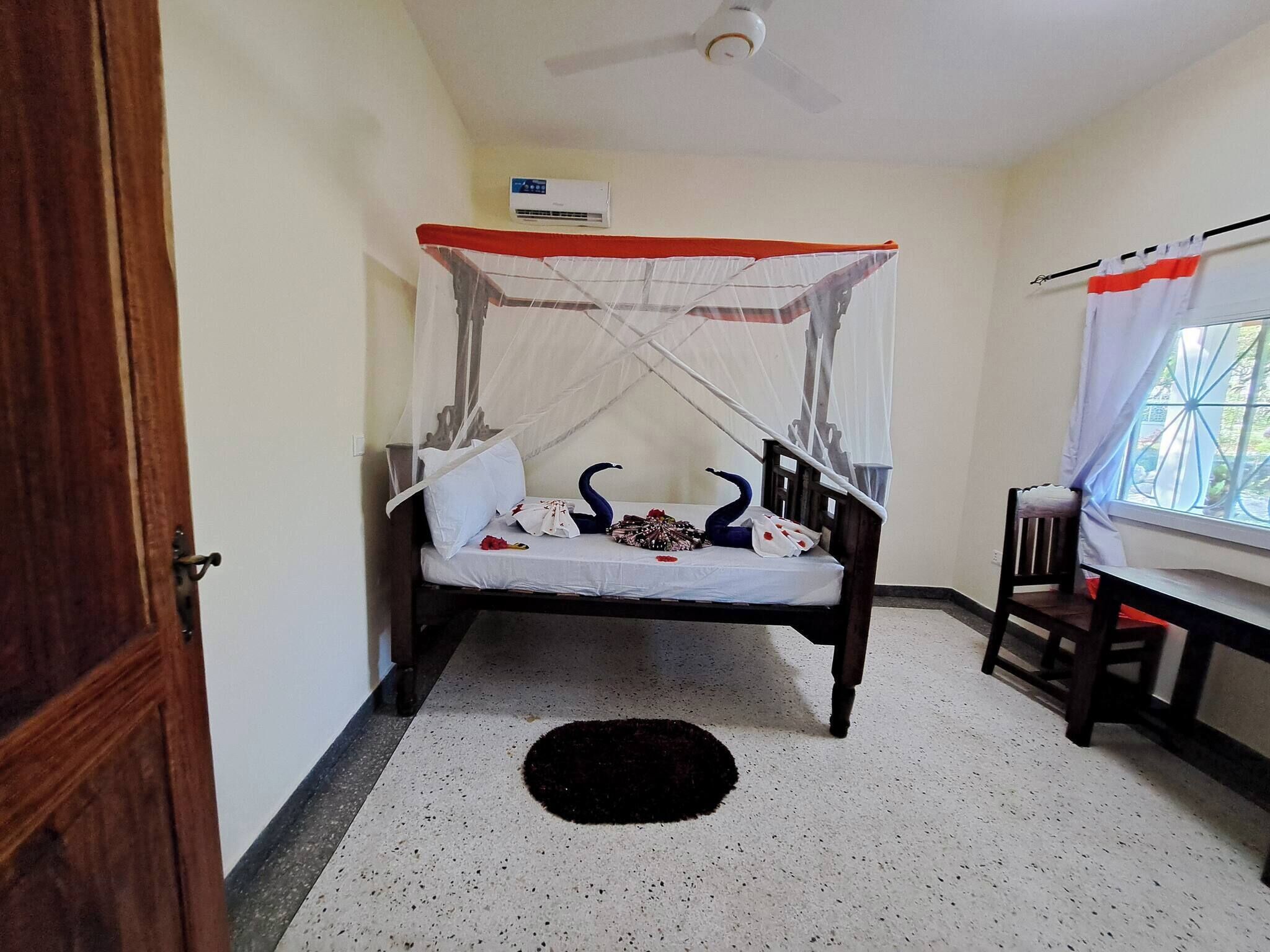 A single bed with a canopy, a ceiling fan, a window with curtains, and a small table with a chair.