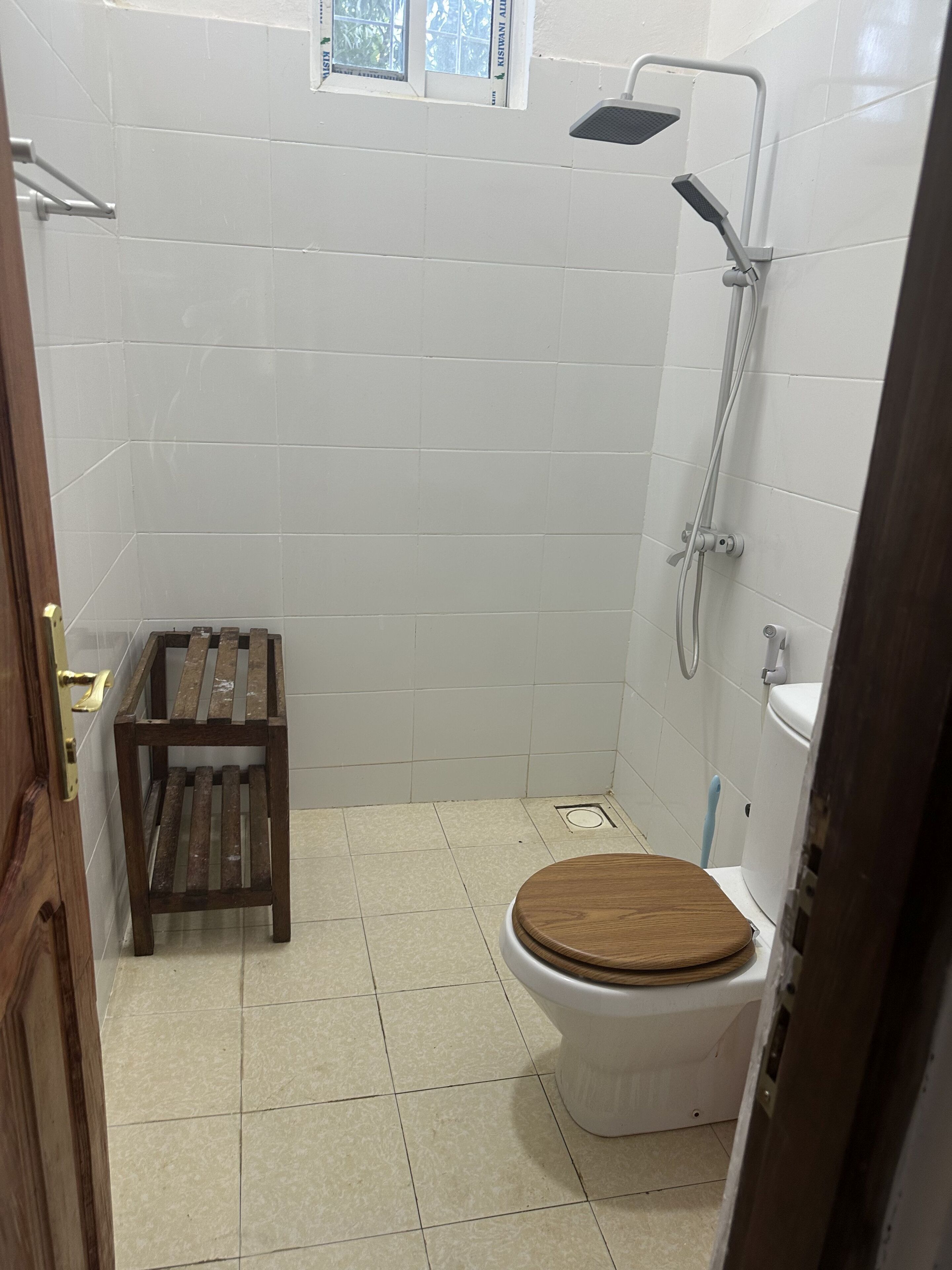 Une petite salle de bain carrelée, équipée de toilettes, d’un tabouret en bois et d’une douche.