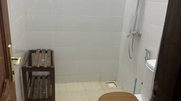 Une petite salle de bain carrelée, équipée de toilettes, d’un tabouret en bois et d’une douche.