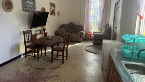 Sala de estar com televisão, sofá, mesa de jantar, cadeiras e área de cozinha com pia e cesto verde.