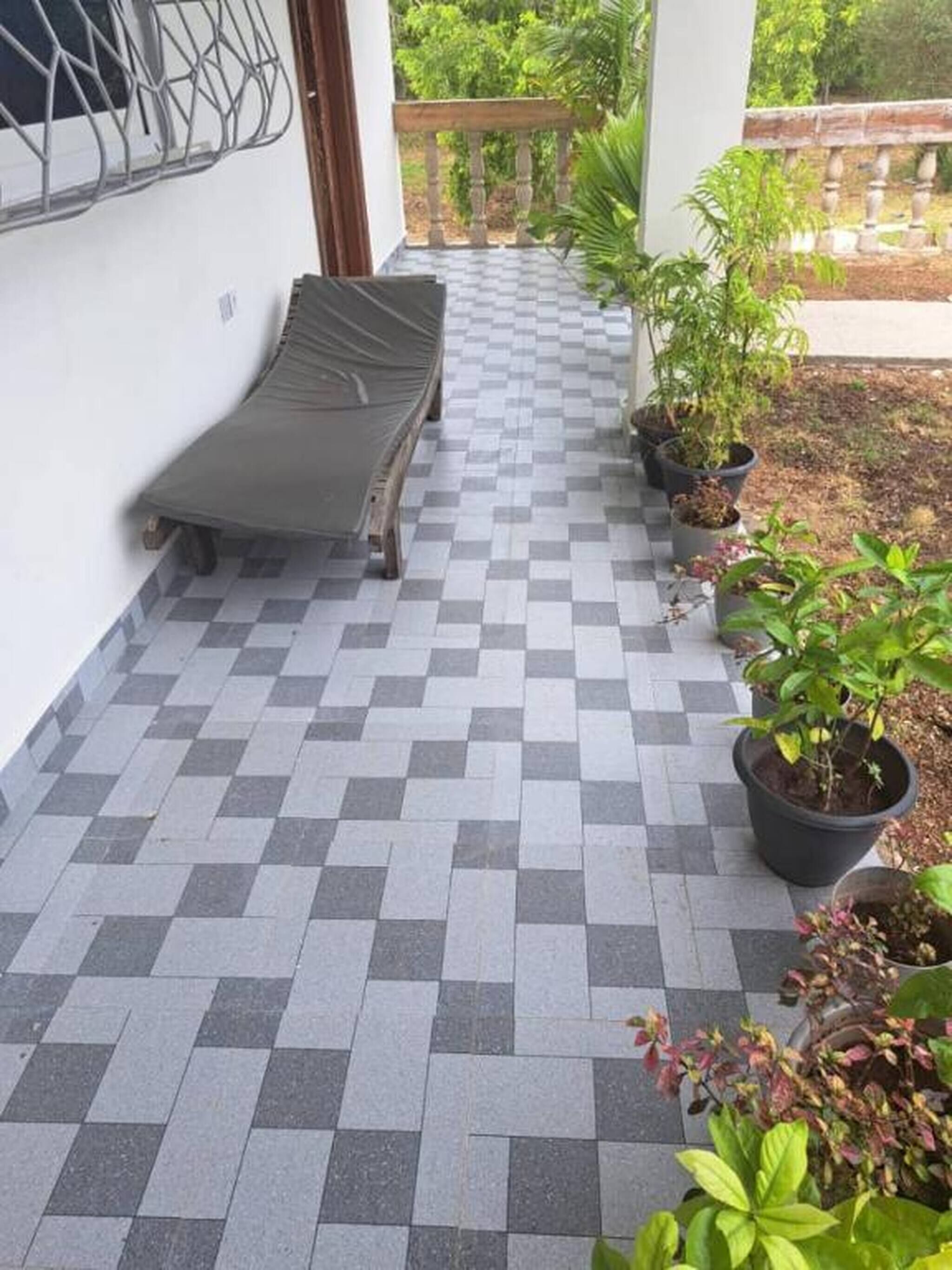 Un balcón con suelo de baldosas a cuadros, una silla de salón, plantas en macetas y vistas al verdor.