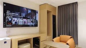 Ein modernes Hotelzimmer mit einem an der Wand befestigten Flachbildfernseher, der einen Film zeigt. Es gibt einen Holztisch mit einem Stuhl, einen kleinen Hocker und einen schwarzen Kühlschrank.