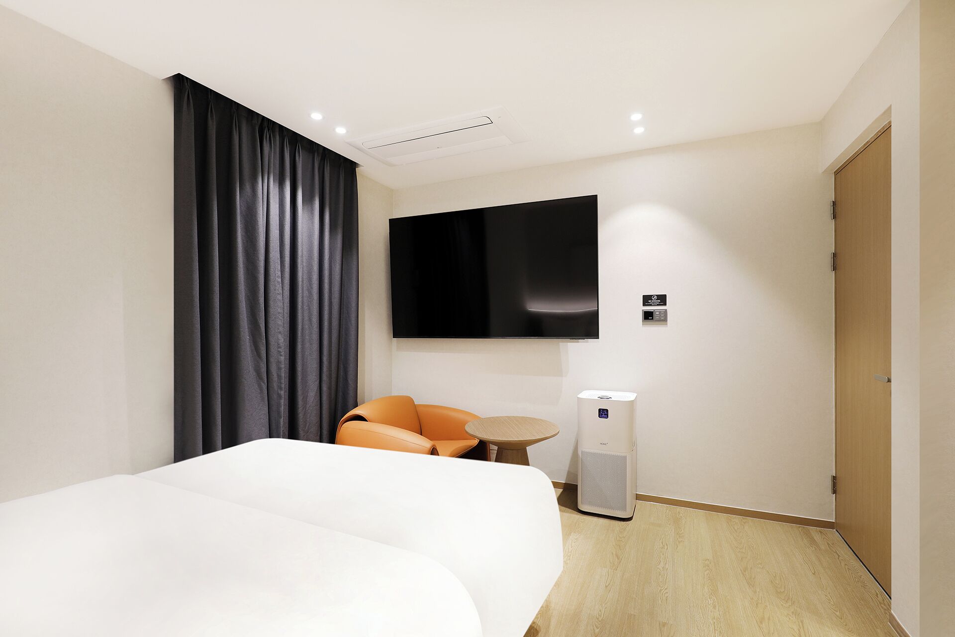 A modern hotel room with a large bed, a flat-screen TV, a small round table, an air purifier, and a door.