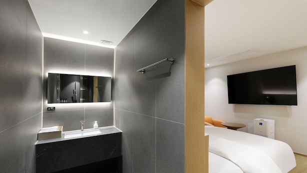 Ein modernes Badezimmer mit einem großen Spiegel, einer Dusche und einem Waschbecken. Im angrenzenden Zimmer befindet sich ein Bett mit einem Fernseher.