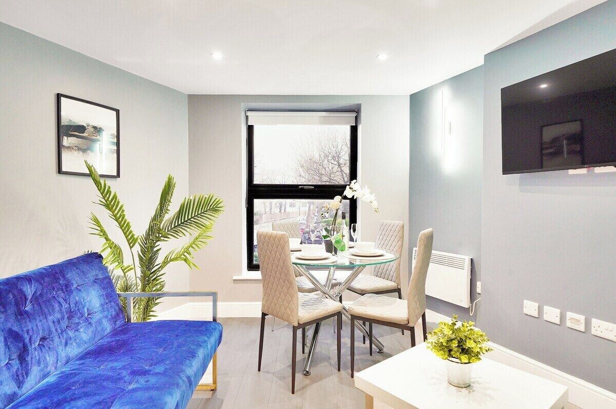 Ein modernes Wohnzimmer mit einem blauen Sofa, einem Glastisch mit Stühlen, einer Topfpflanze und einem großen Fenster.