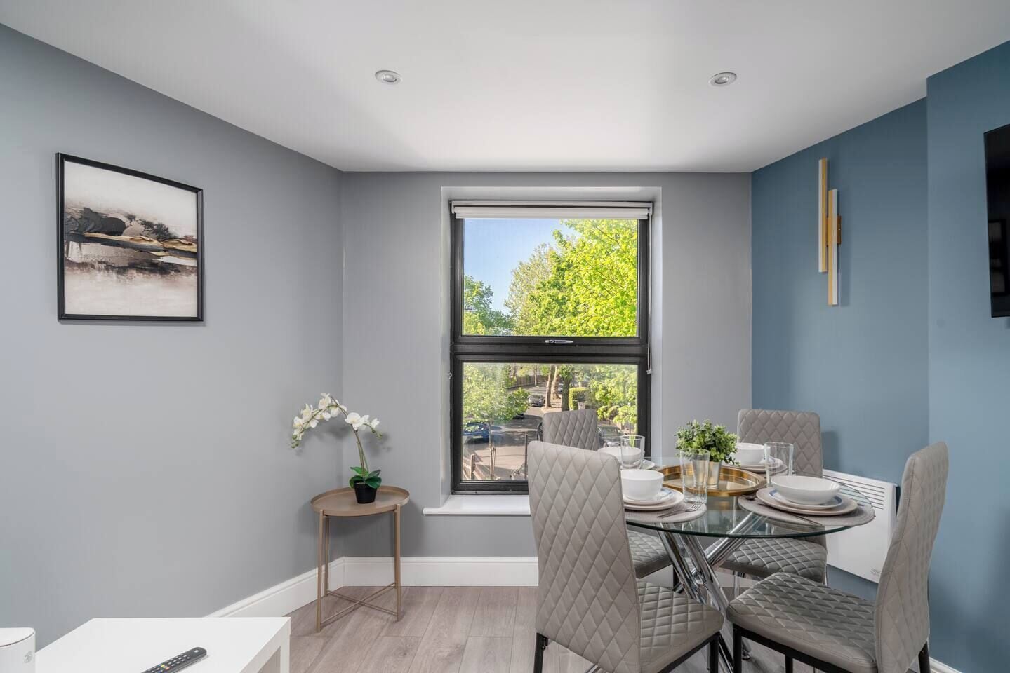 A dining area with a glass table, cushioned chairs, a potted plant, and a framed picture on the wall.