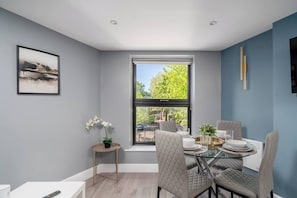 A dining area with a glass table, cushioned chairs, a potted plant, and a framed picture on the wall. - Chic 1-Bed Apartment in Ealing (London)