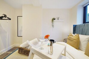 A hotel room with a bed, a bedside table with a vase and a glass, a hanging plant, and a window with curtains.