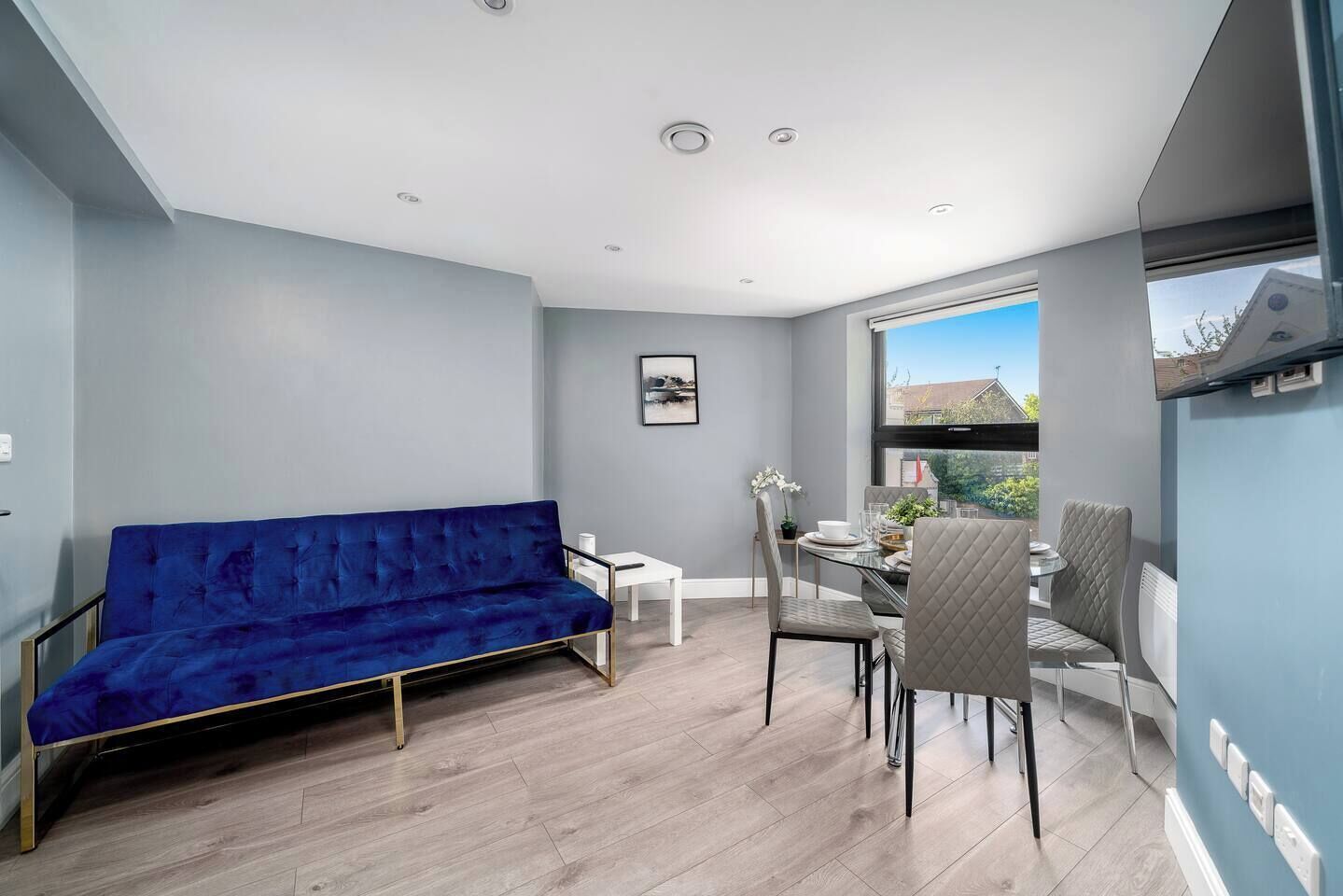 A modern living space with a blue sofa, a dining table with chairs, and a large window offering a view of the outdoors.
