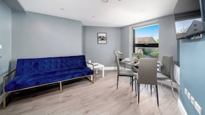 Ein modernes Wohnzimmer mit einem blauen Sofa, einem Esstisch mit Stühlen und einem großen Fenster mit Blick ins Freie.