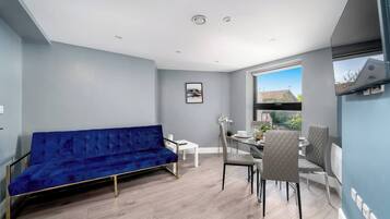 A modern living space with a blue sofa, a dining table with chairs, and a large window offering a view of the outdoors.