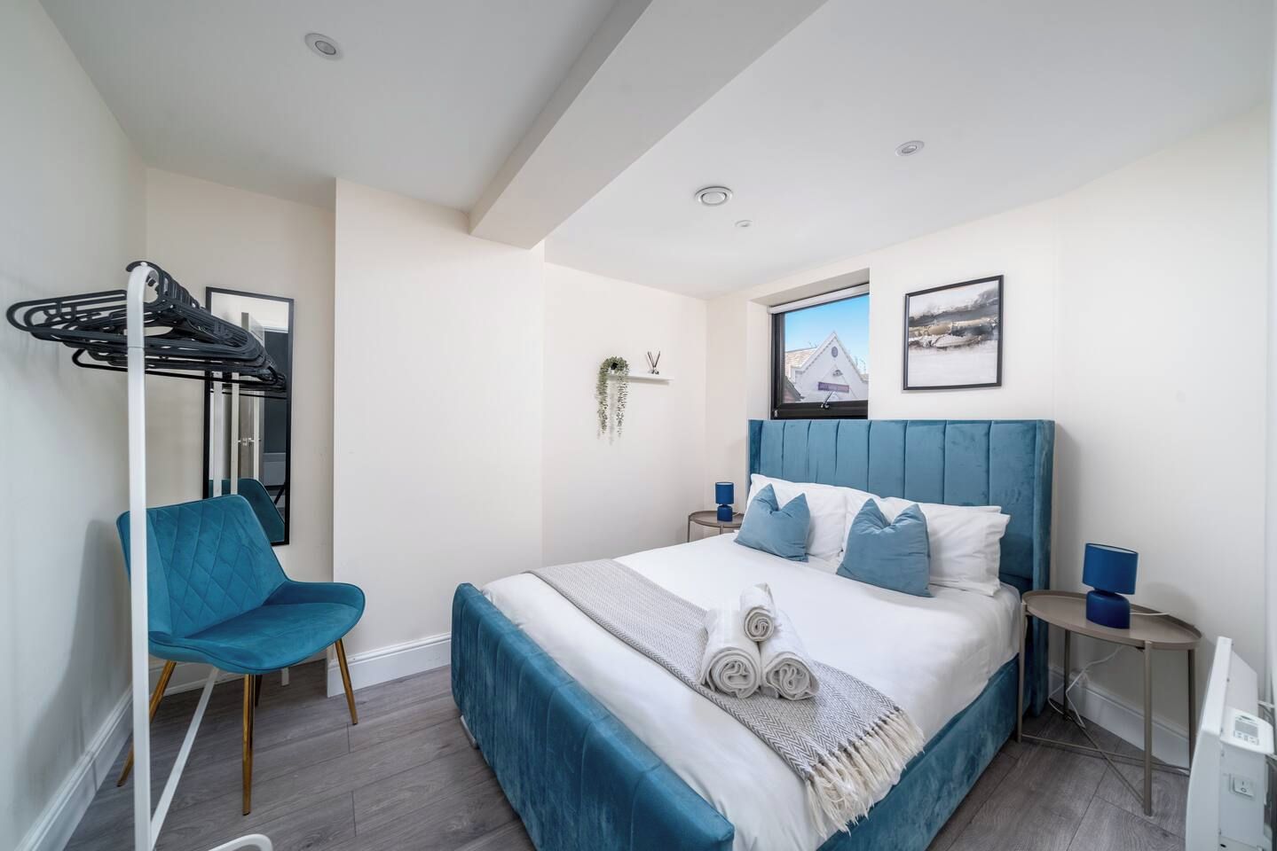 A hotel room with a bed, a blue chair, a small table, a wall-mounted towel rack, and a window with a view.
