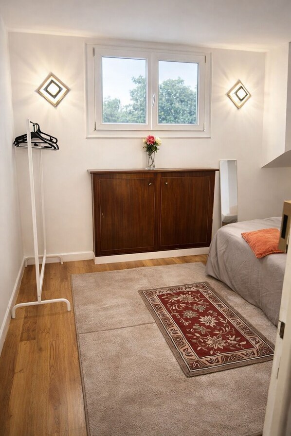 Une chambre à coucher comprenant un lit, une armoire en bois, un portant à vêtements, une fenêtre et un tapis décoratif.