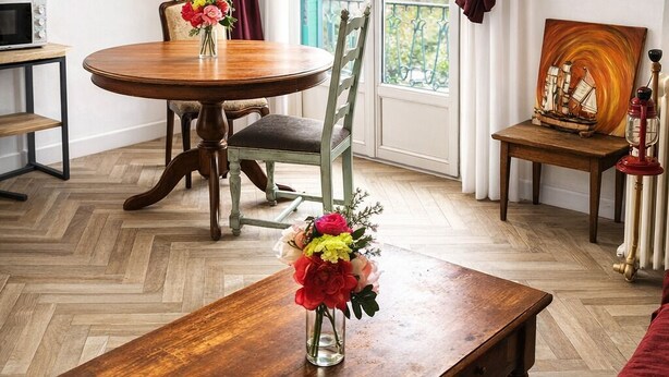 Ein Zimmer mit Holzboden, einem runden Esstisch mit Stühlen, einer Couch, einem kleinen Tisch mit Blumen und einem Fenster mit Vorhängen.