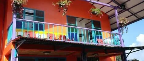 A two-story building with a vibrant orange facade, a balcony with blue railings, and hanging plants.