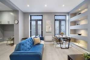 A modern living room with a blue sofa, a dining table with chairs, and a kitchen area with cabinets and a clock. - Stunning 1 Bedroom Apartment (London)