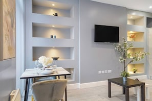 A modern living room with a dining area, a flat-screen TV mounted on the wall, and built-in shelving with decorative items. - Stunning 1 Bedroom Apartment (London)