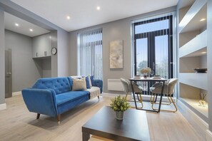 A modern living room with a blue sofa, a dining table with chairs, a potted plant, and a large window with blinds. - Stunning 1 Bedroom Apartment (London)
