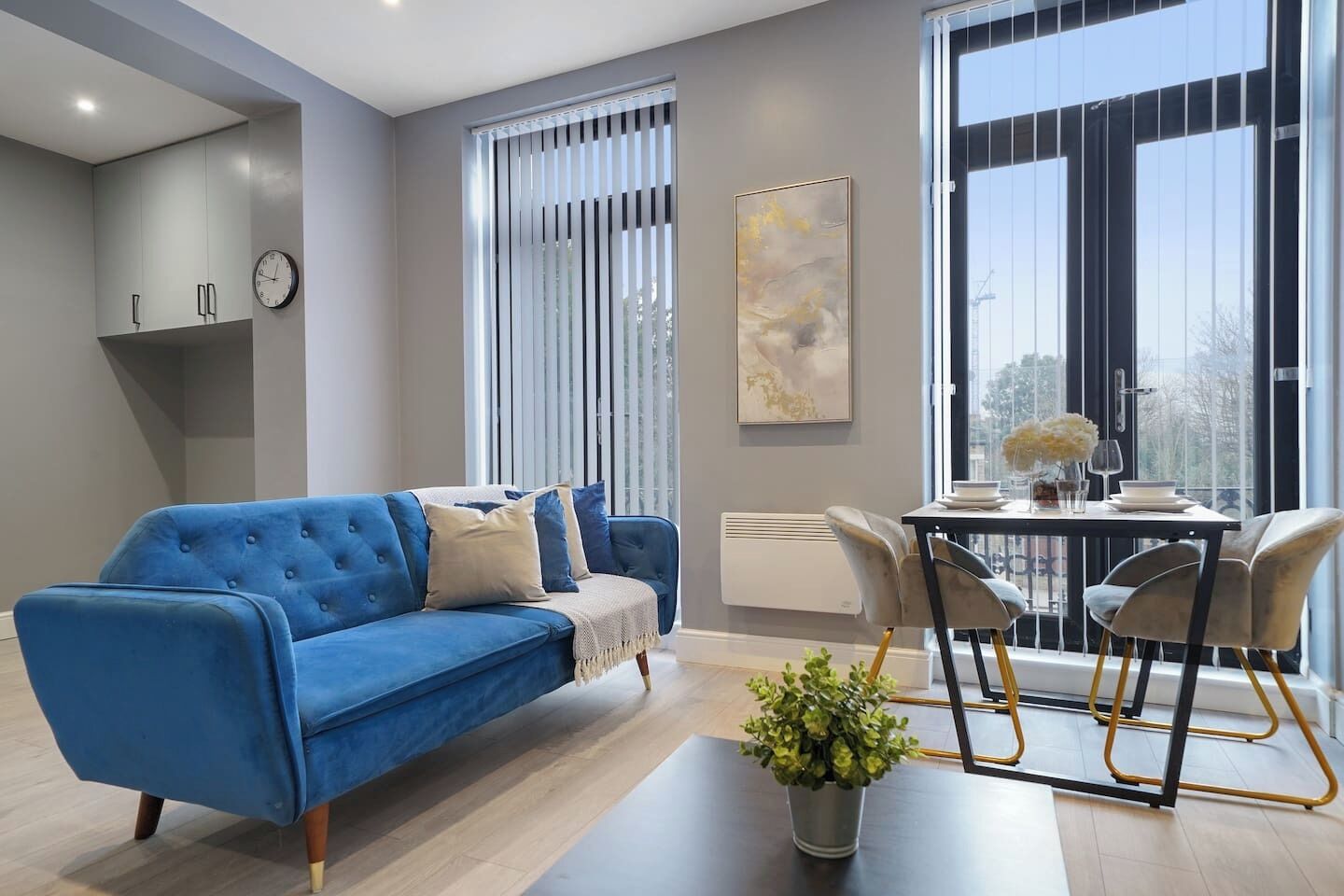 A modern living room with a blue sofa, a dining table with chairs, a potted plant, and a large window with blinds.