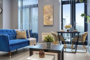 A modern living room with a blue sofa, a coffee table, a dining table with chairs, a potted plant, and a large window with blinds. - Stunning 1 Bedroom Apartment (London)