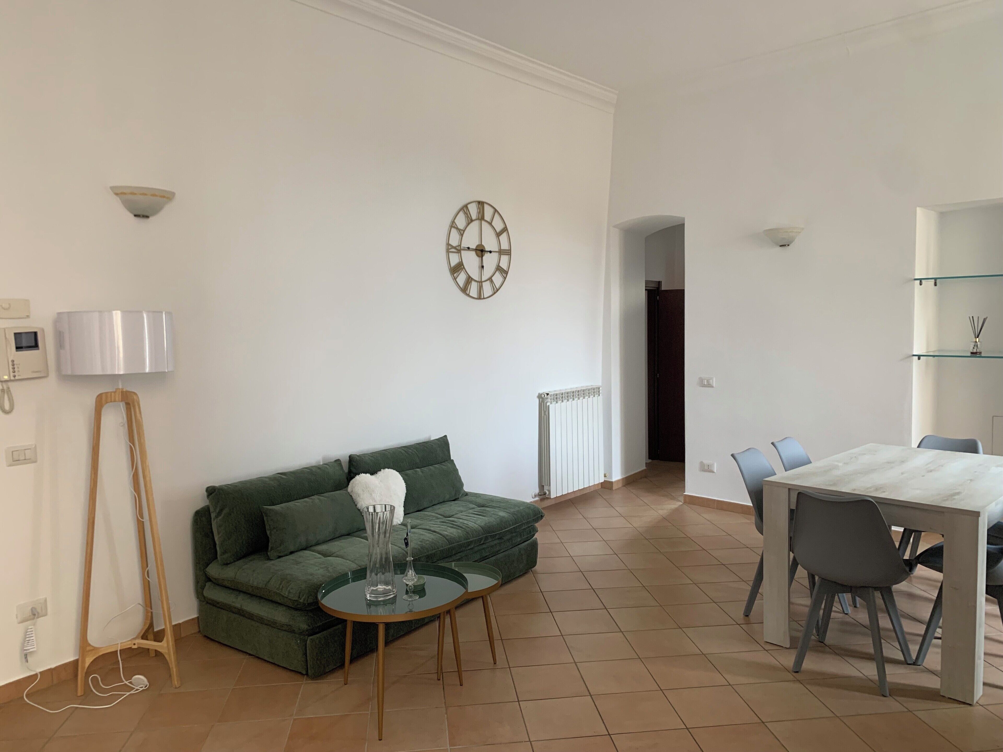 A modern living room with a green sofa, a round glass coffee table, a white lamp, a wall clock, and a dining area with a marble table and chairs.