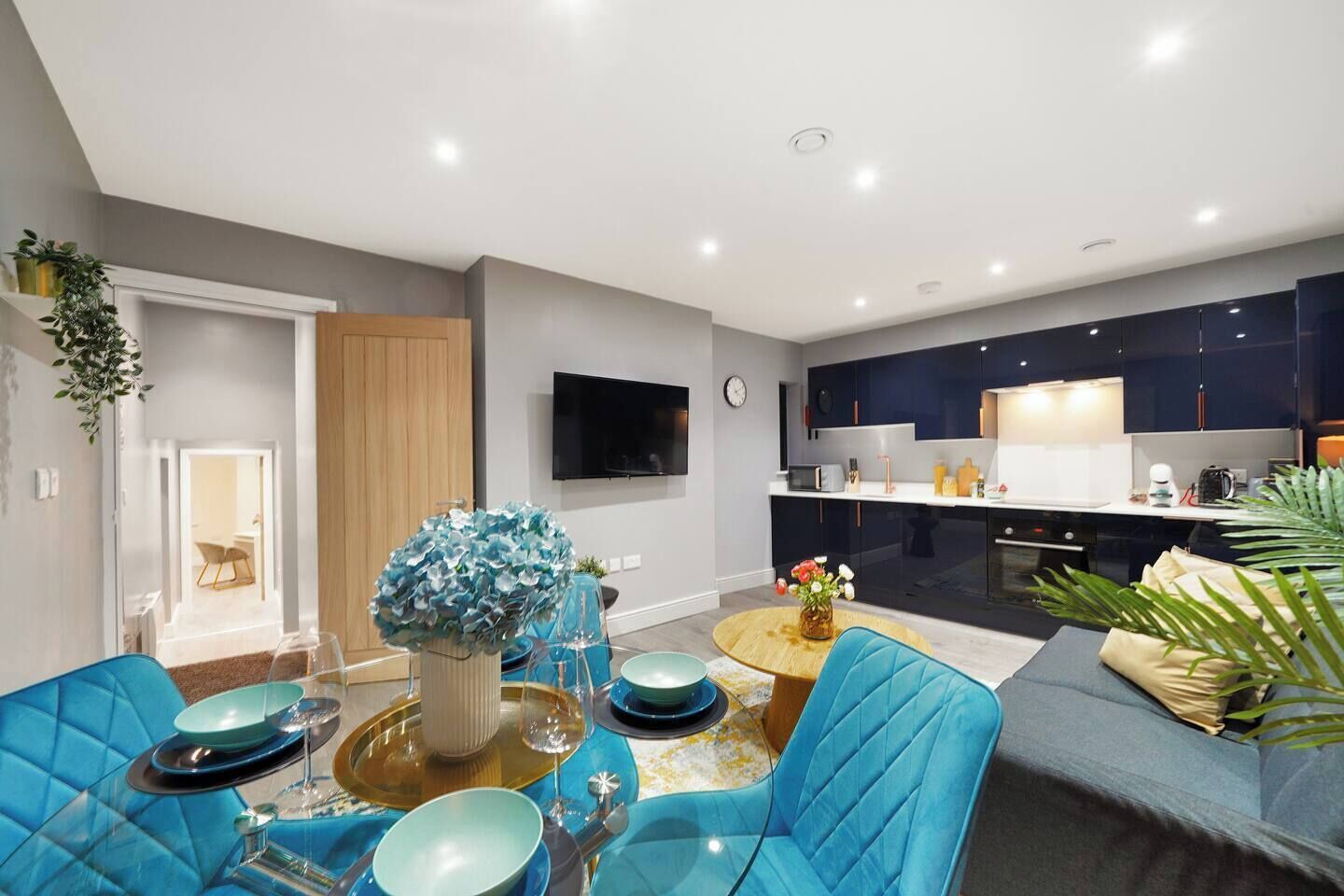 A modern living room with a dining area, a kitchen with dark cabinets, and a flat-screen TV mounted on the wall.