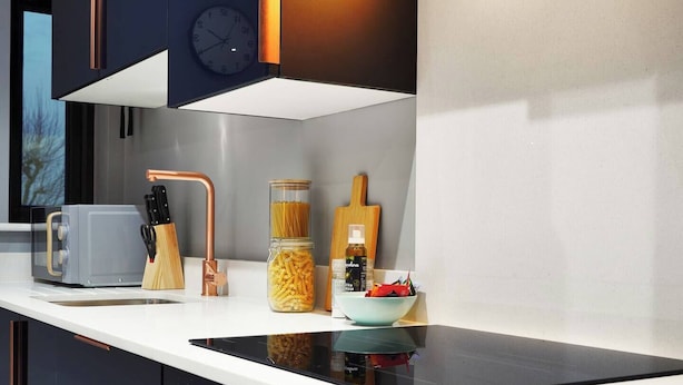 Una cocina moderna con horno empotrado, cafetera, bloque de cuchillos, tarro de pasta, tabla de cortar y un bol de fruta.