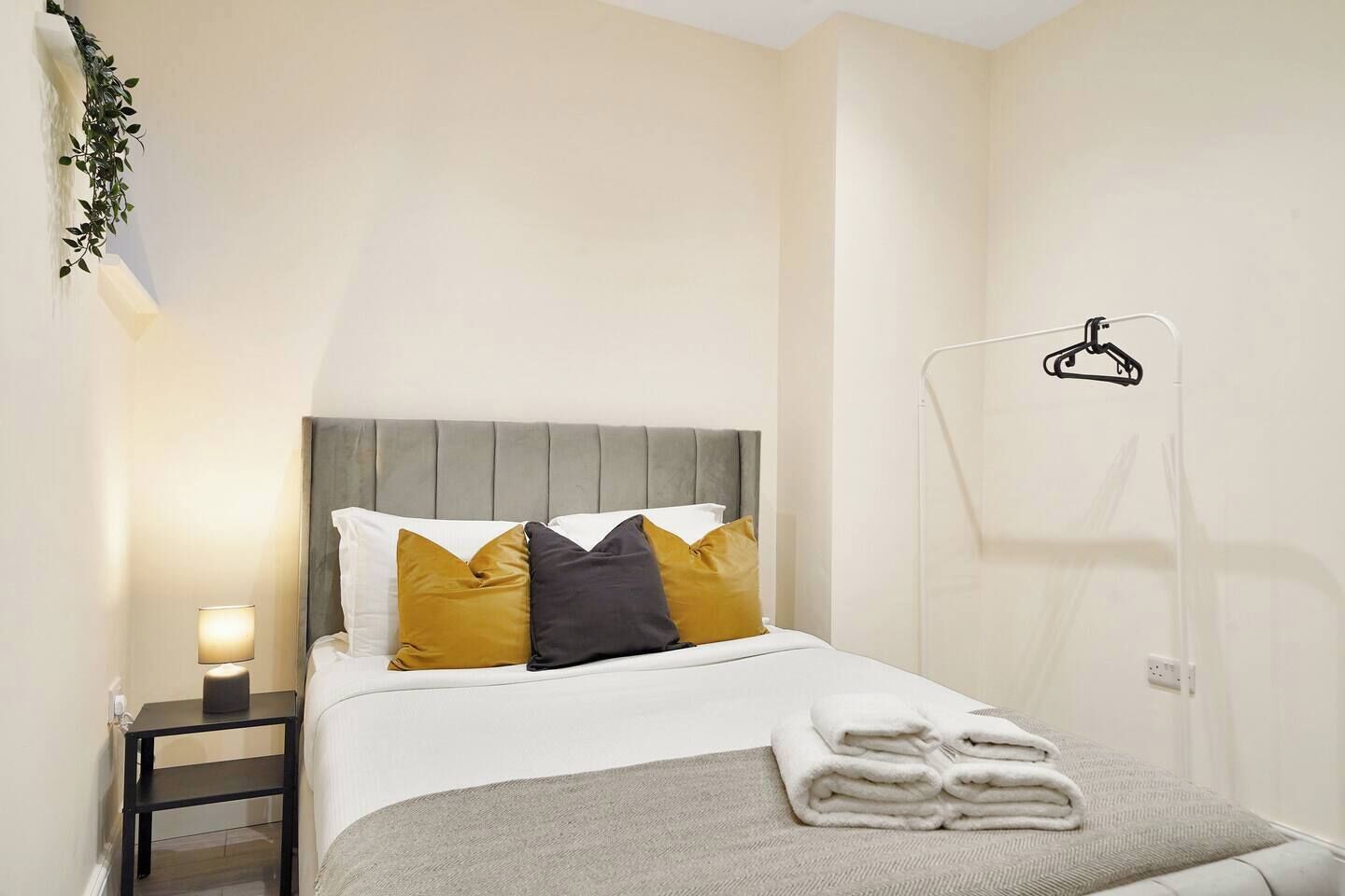 A neatly made bed with a grey headboard, a bedside table with a lamp, and a towel rack.