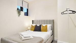 Un dormitorio con cama, almohadas, una lámpara de noche, una planta colgante y un perchero.