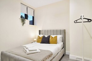 A bedroom with a bed, pillows, a bedside lamp, a hanging plant, and a coat rack. - Modern & Cozy 3-Bed Apartment (London)