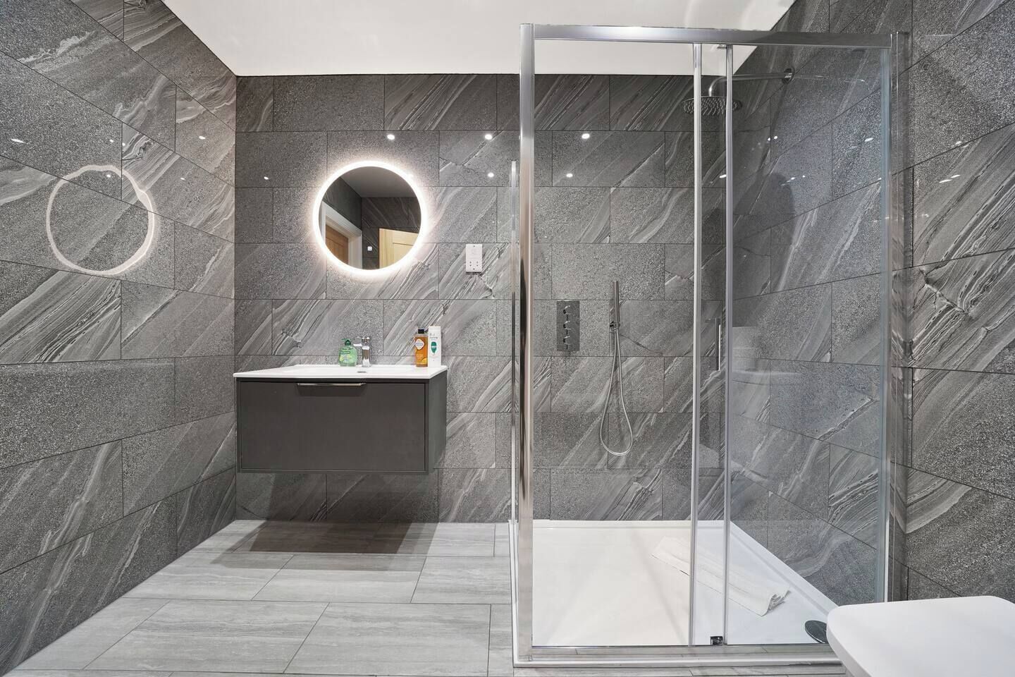 Un bagno moderno con cabina doccia in vetro, uno specchio rotondo e una parete rivestita in marmo.