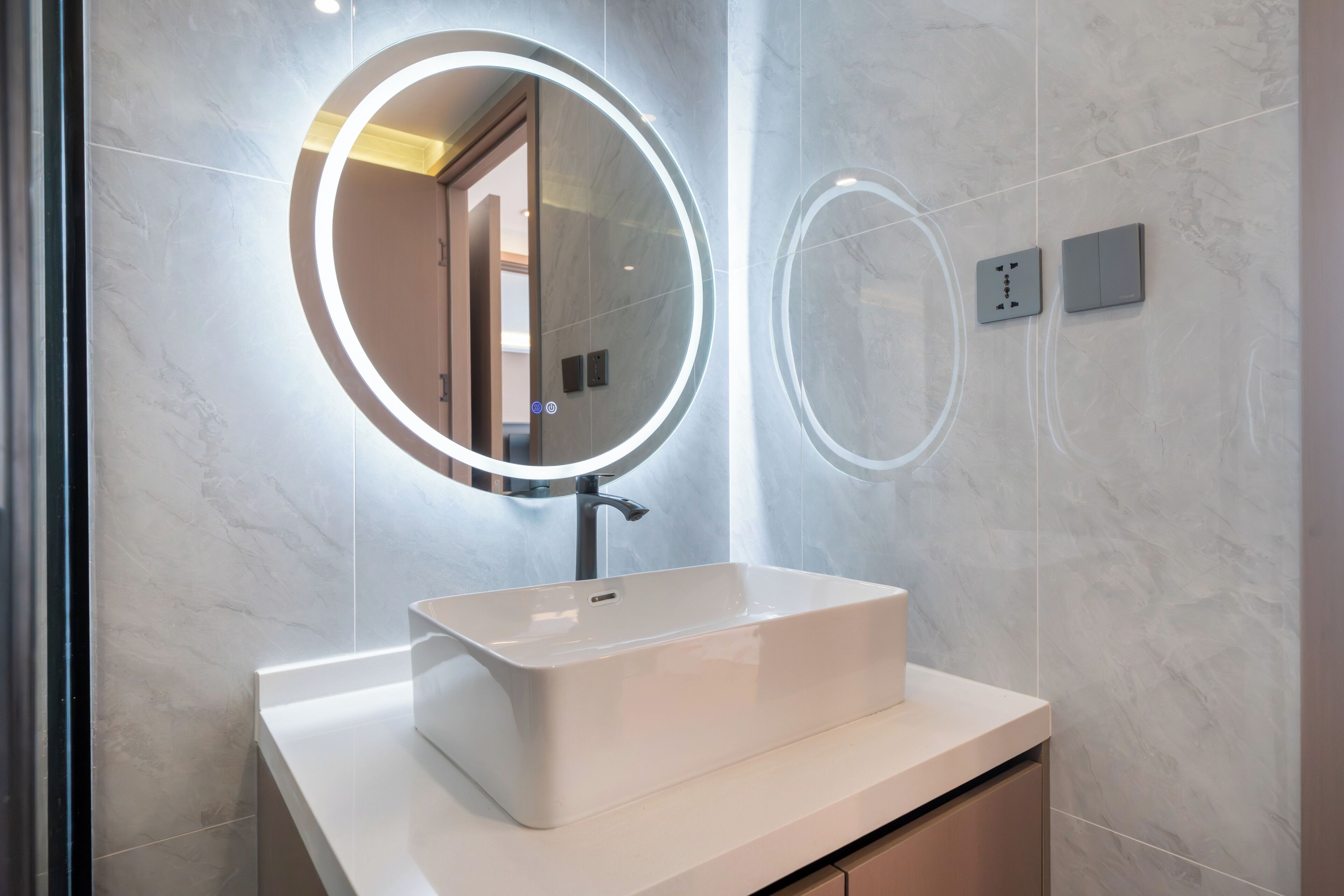 A bathroom with a white sink, a round mirror with lights, and a marble wall.