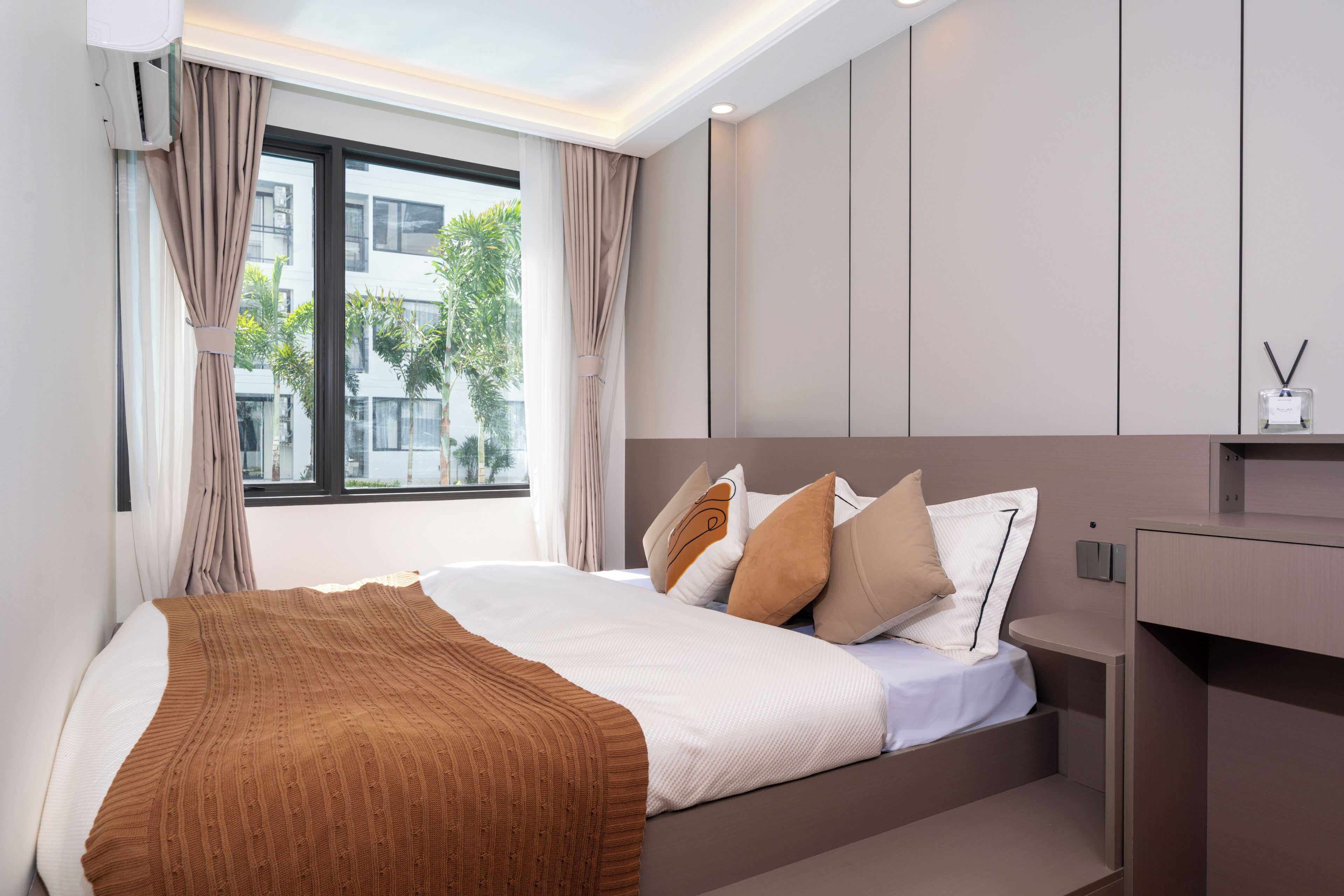 A modern hotel room with a bed, curtains, a window with a view of buildings and trees, and a bedside table with a lamp.