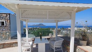 A covered outdoor seating area with a table and two chairs, overlooking a scenic view of the sea and mountains.