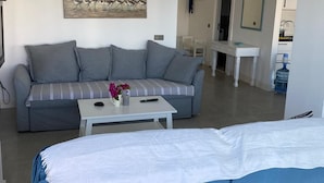 A modern living room with a grey sofa, a white coffee table, and a blue sofa. There is a painting on the wall and a kitchen area visible through an open door.