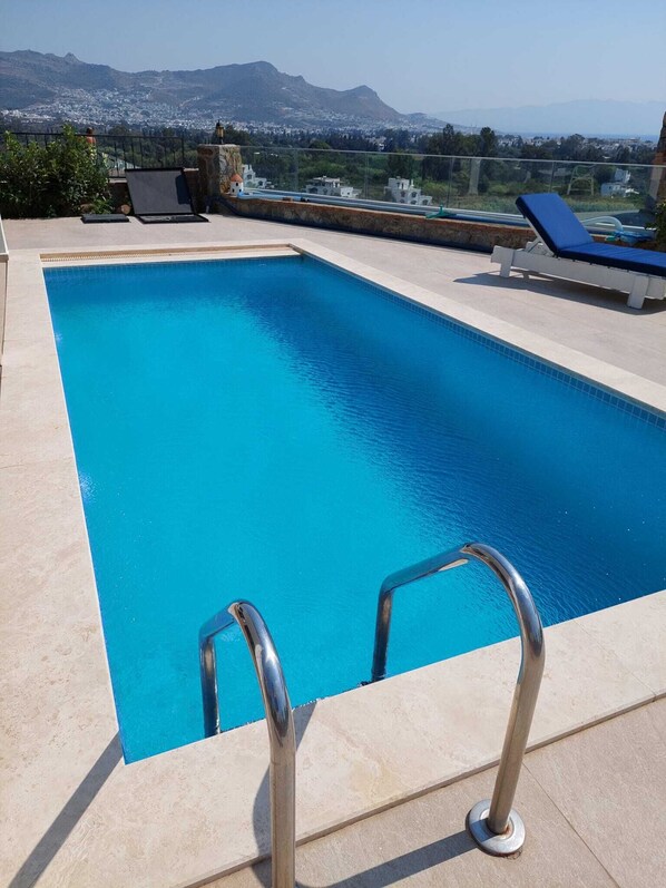 A rectangular swimming pool with clear blue water, surrounded by a tiled deck. There are metal pool ladders at the shallow end. In the background, there are mountains and a view of a town. - Panoramic view - modern apartment with large private pool area, beach 1km (Bodrum - Peksimet)