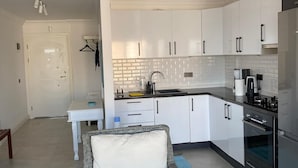 A modern kitchen with white cabinets, a black countertop, and stainless steel appliances. There is a dining area with a wicker chair and a table.