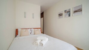 A neatly made bed with white linens and rolled towels, a wooden headboard, and three framed pictures on the wall. - Cosy 2 Bed Flat in Naujamiestis (Luton)