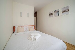 Ein ordentlich bezogenes Bett mit weißen Leintüchern und aufgerollten Handtüchern, ein hölzerner Kopfteil und drei gerahmte Bilder an der Wand.