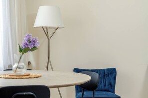 Pyöreä pöytä, jonka päällä on maljakko täynnä violetteja kukkia, moderni lamppu ja sinisellä kankaalla verhoiltu tuoli huoneessa, jossa on vaaleat seinät.