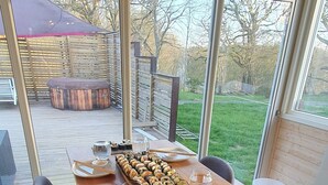 Ein Essbereich mit einem Holztisch, gedeckt für zwei Personen, ein großes Fenster mit Blick ins Freie und eine Holzterrasse, die durch das Fenster sichtbar ist.
