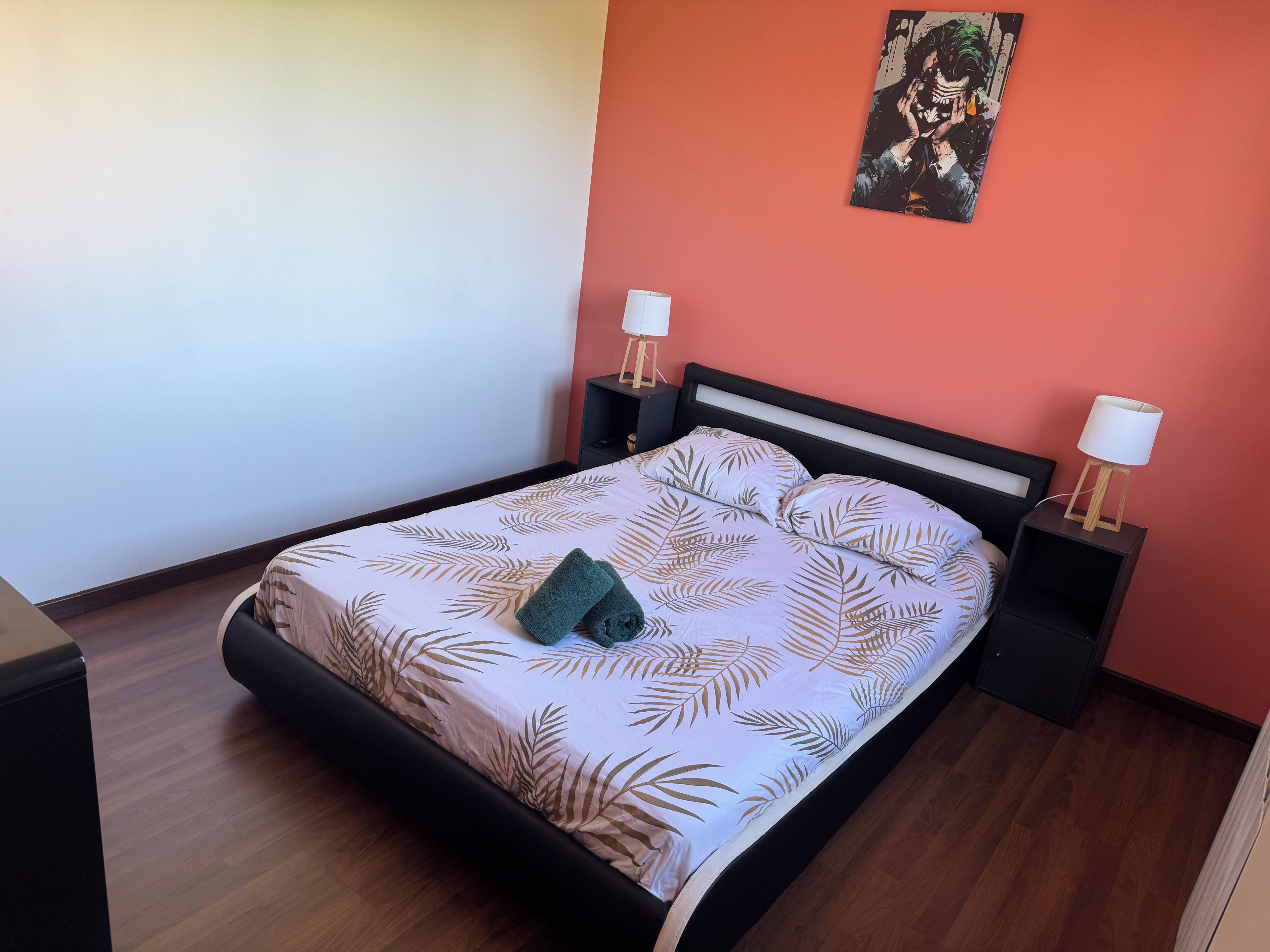 Un lit bien fait, recouvert d’une couvre-lit à motifs de feuilles, deux tables de chevet avec des lampes et un seul tableau au mur.
