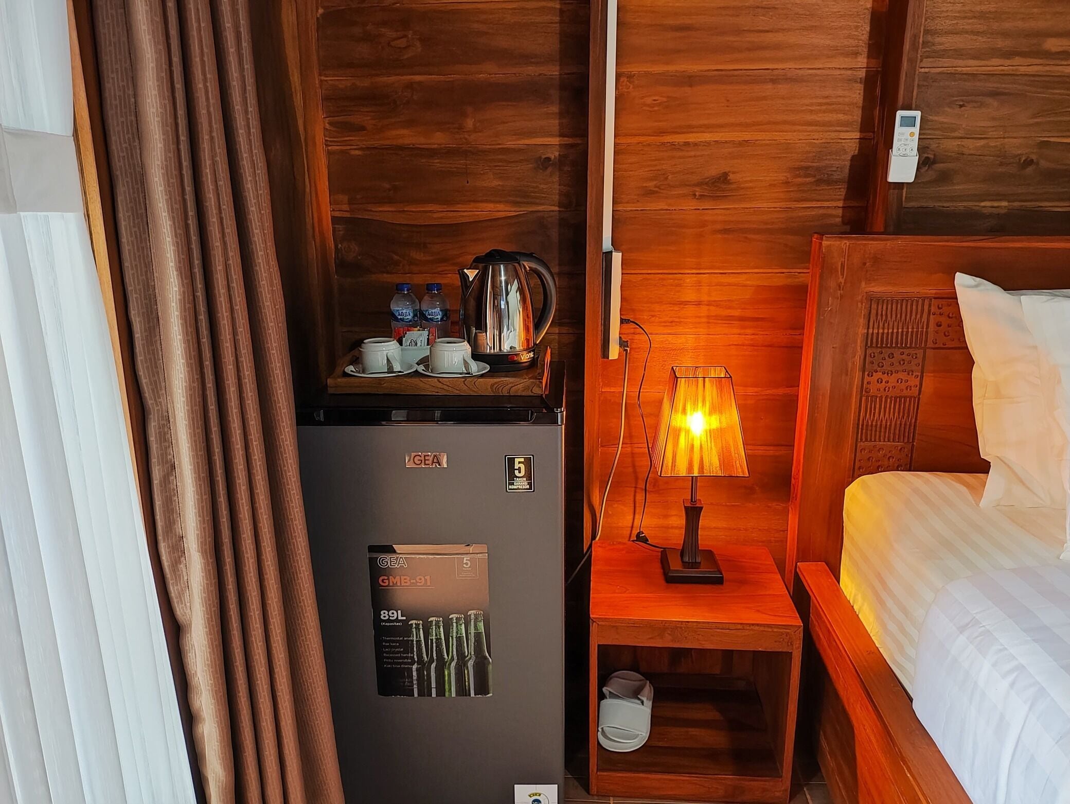 A hotel room with a GEA refrigerator, a wooden nightstand with a lamp, a bed, and a wooden headboard.