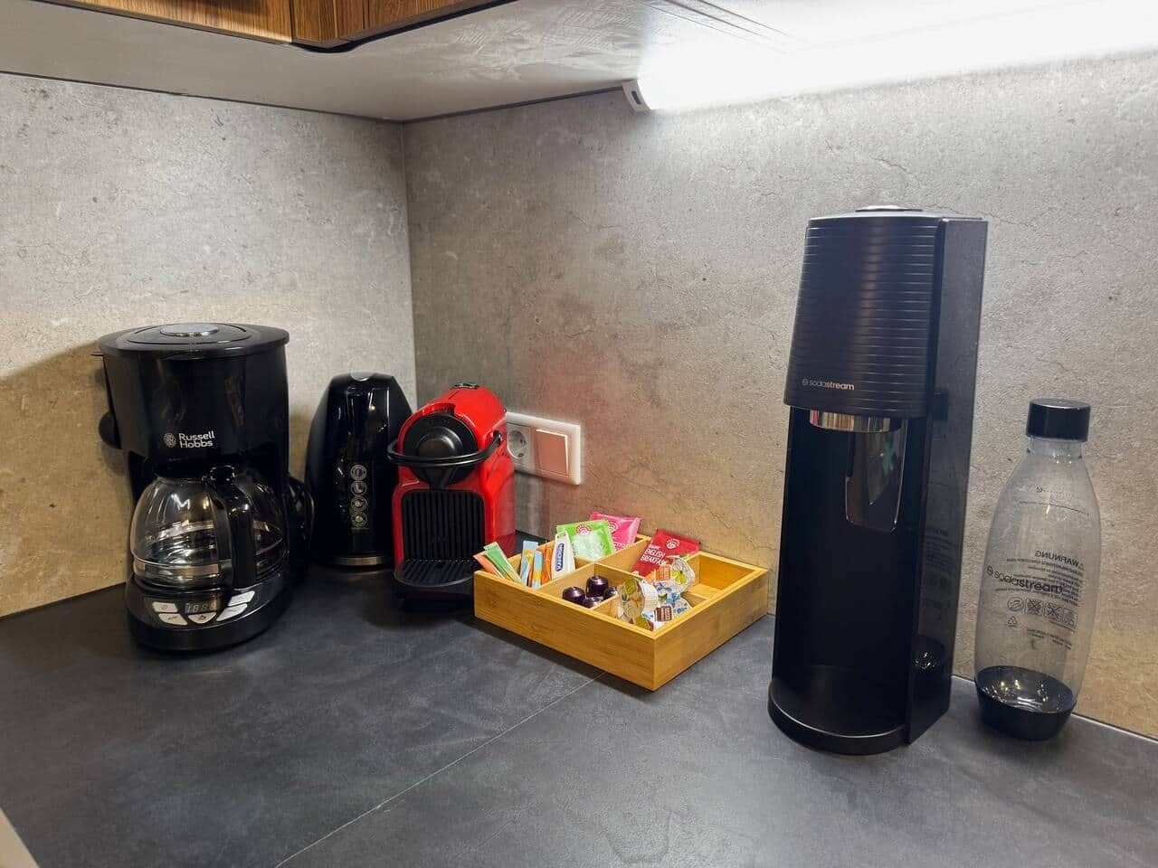 En köksbänk med en kaffebryggare, en röd kaffemaskin, en svart kaffemaskin, en trälåda med snacks och en vattenflaska.