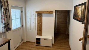 Een kleine, goed verlichte kamer met houten vloer, een wit bureau, een klein kastje, een raam met gordijnen en een lijst met een afbeelding aan de muur.