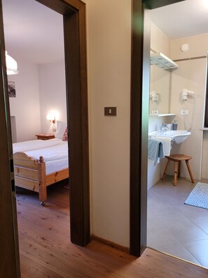 A hotel room with a wooden bed, a bedside lamp, a small stool, a bathroom with a sink and mirror, and a tiled floor. - Apartment 'Casa Rustica App Bulaccia' with Mountain View, Shared Garden & Balcony (Sankt Ulrich)